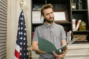 Business professional standing confidently in office holding documents near American flag.